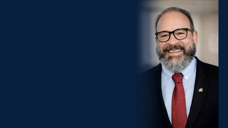 Dr. Maurício Ribas, advogado, sorrindo, de óculos, barba grisalha, terno escuro e gravata vermelha, à direita de um fundo azul-marinho